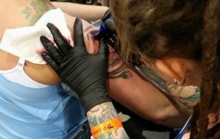 A person with tattoos and dreadlocks is giving a shoulder tattoo to a woman with blonde hair and colorful dye. The woman is seated on a chair in a tattoo parlor.