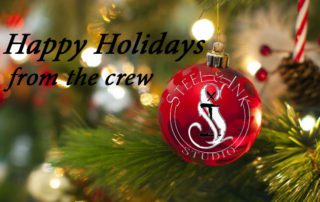 A red holiday ornament with the text "STEEL & INK STUDIO" hangs on a Christmas tree. The words "Happy Holidays from the crew" are written in black on the left side of the image.