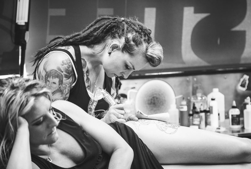 Black and white photo of a woman getting a tattoo on her leg.